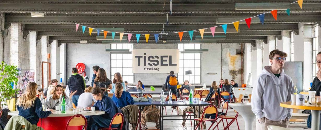 intérieur de l'ancienne usine Tissel à Roubaix, où l'on voit plusieurs personnes attablées par petits groupes, et des fanions colorés qui décorent